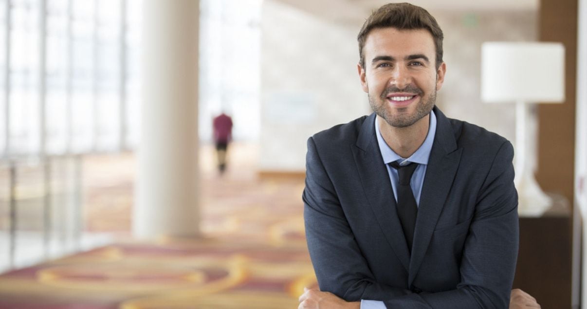 Hombre joven adulto profesional de la hoteleria especializado en revenue management sonrie mientras da la bienvenida a los huespedes en el lobby de un hotel de 5 estrellas