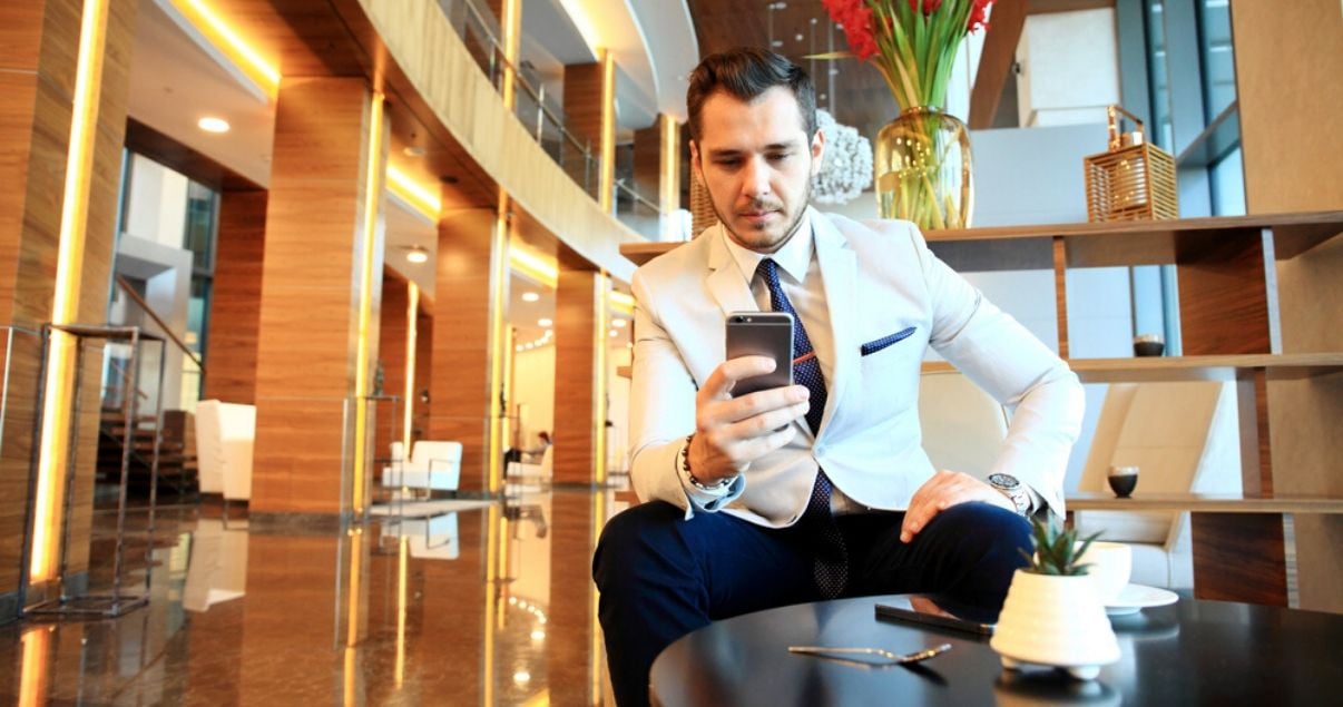 Hombre joven adulta vestido con saco balnco corbata y pantalon azules sentado en el lobby de un hotel mirando la pantalla de su celular en el que analiza reportes de revenue management para entender como aumentar la 