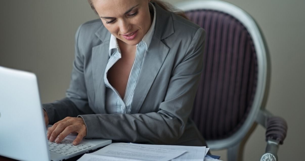 Mujer joven adulta de tez blanca vestida con traje gris y camisa a rayas celestes sentada frente a laptop blanca y reportes de revenue management