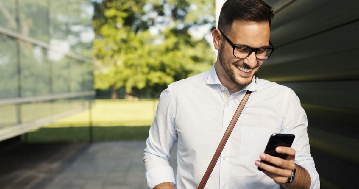 Hombre joven adulto vestido con camisa y anteojos de marco grueso negros mira la pantalla de su celular con alegria porque acaba de reservar una habitacion de hotel para sus vacaciones usando bitcoins como forma de pago