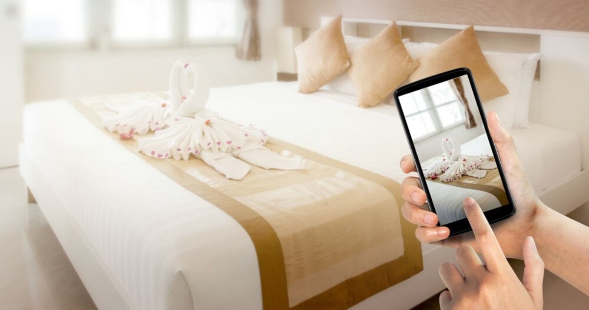 Manos de mujer joven adulta parada frente a la cama de dos plazas de un hotel mientras saca una fotografia con su celular para web para hoteles 