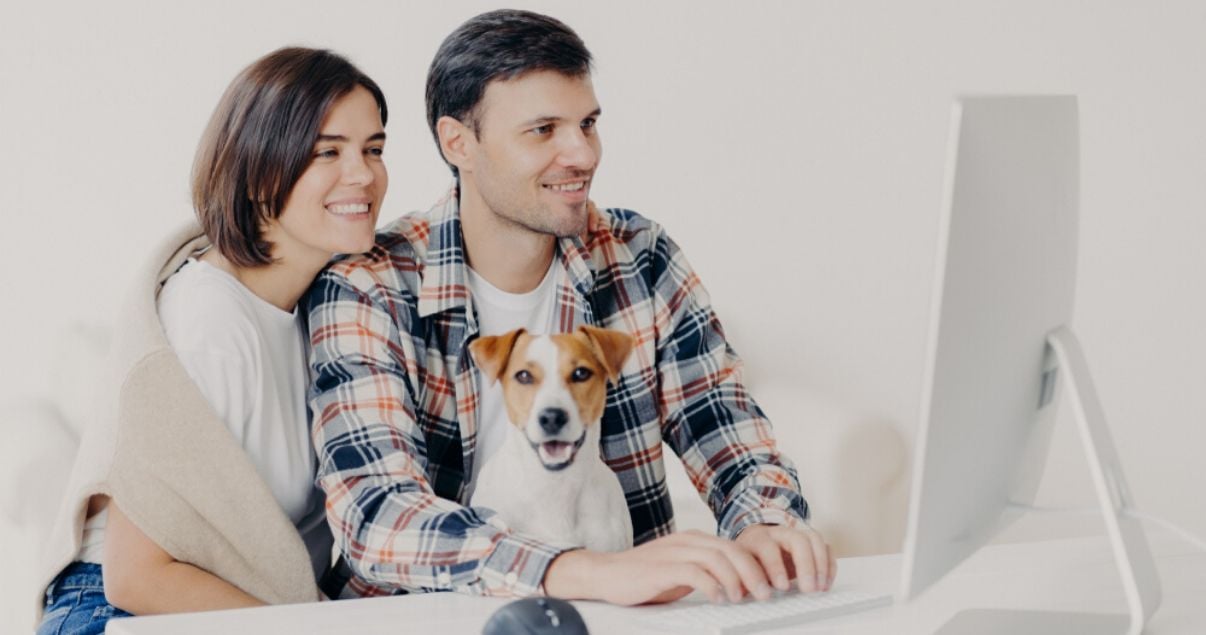 Primer plano de una pareja de hombre y mujer jovenes adultos sentados frente al monitor blanco de su computadora junto a su pequeno perro mientras dejan resenas sobre sus experiencias en hoteles pet friendly en las u