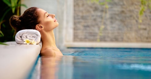 Primer plano de una mujer joven adulta de cabello castano posando para produccion de un articulo sobre hoteles con piscina mostrandose muy relajada en el agua con la cabeza recostada sobre una toalla blanca apoyada e