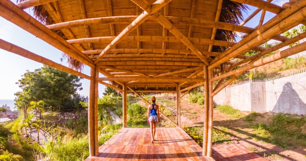 Plano general de una mujer joven adulta con cabello castano recogido vestida en shorts y musculosa caminando sobre una de las cabanas de madera que caracterizan a los hoteles sustentables