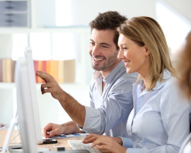 Hombre y mujer jovenes adultos parte del personal de un hotel trabajando sonriendo frente al monitor de una computadora de escritorio mientras gestionan reservas facilmente a traves de un channel manager